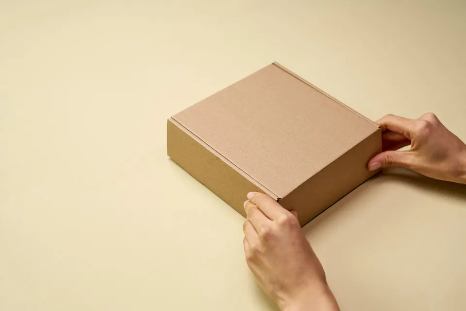 Mãos abrindo uma caixa de papelão marrom sobre uma superfície clara