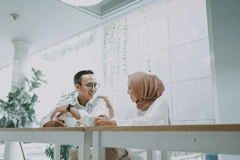 Pasangan muda, perempuan berhijab, tertawa berdua di meja kafe dengan dua cangkir