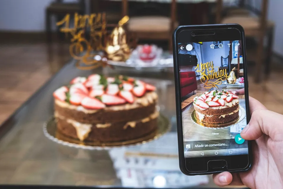A hand holding a phone that shows a photo of a strawberry birthday cake, with the real cake and a gold “Happy Birthday” topper behind it