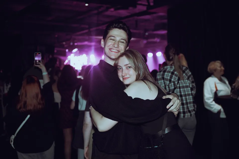 A smiling young couple hugging at a concert, with purple stage lights and the crowd behind them