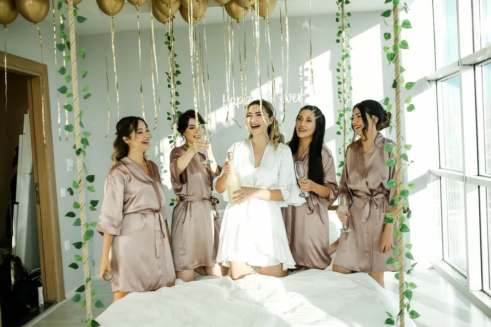 A bride and her bridesmaids in matching robes laugh and raise their glasses in a bright room