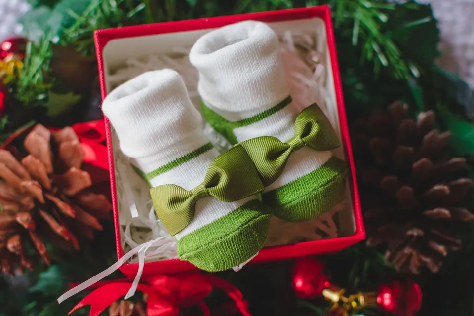 A pair of white baby socks with green bows inside a red Christmas gift box, among pine cones and ornaments