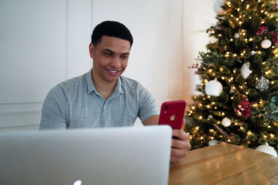 A young man smiles at his phone at a table next to a lit Christmas tree