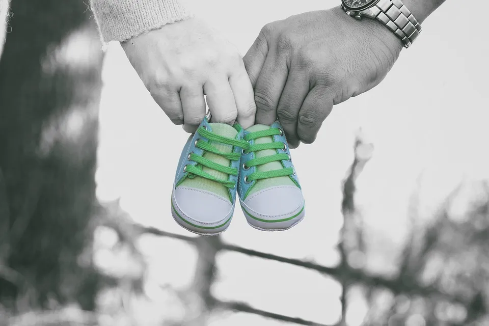 Two hands hold a tiny pair of green baby sneakers by the laces