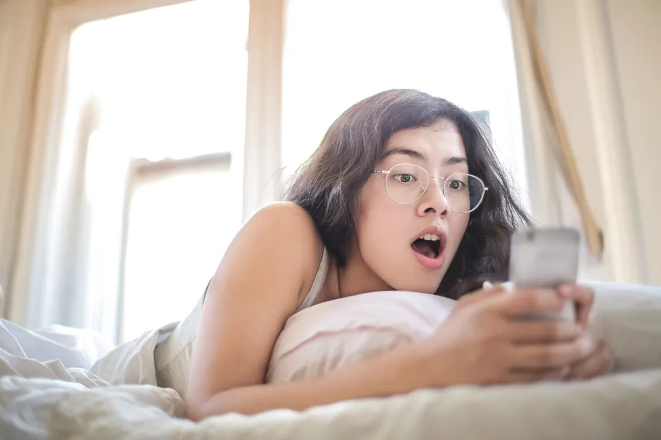 A young woman in glasses lying in bed looks at her phone with her mouth open in surprise