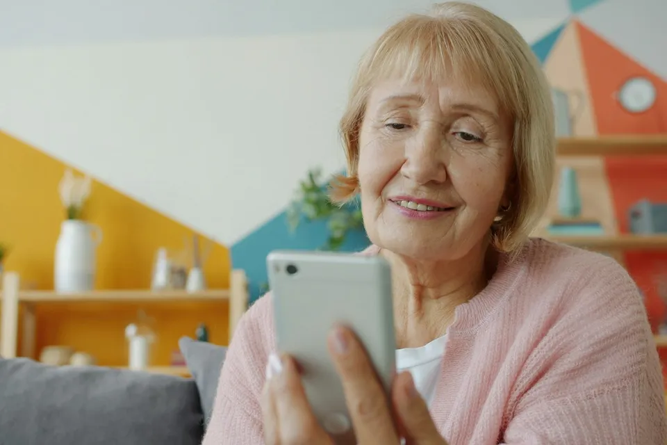 Une femme âgée sourit en regardant l'écran de son téléphone, assise sur son canapé