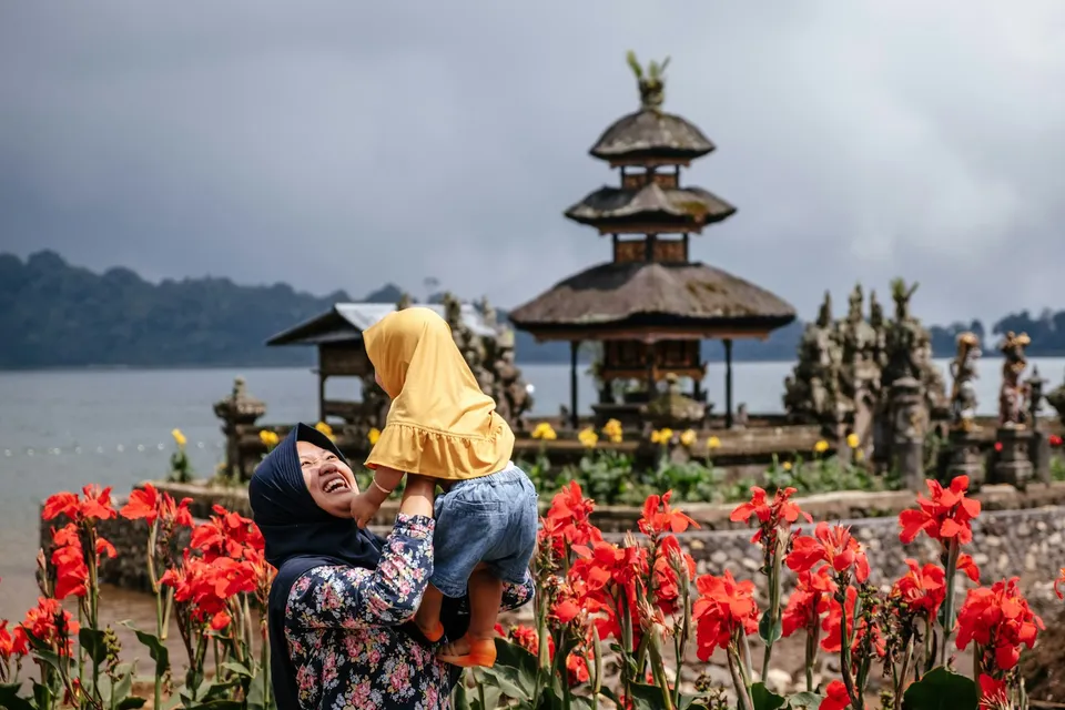 Seorang ibu berhijab menggendong anak perempuannya sambil tertawa di depan Pura Ulun Danu Beratan, Bali