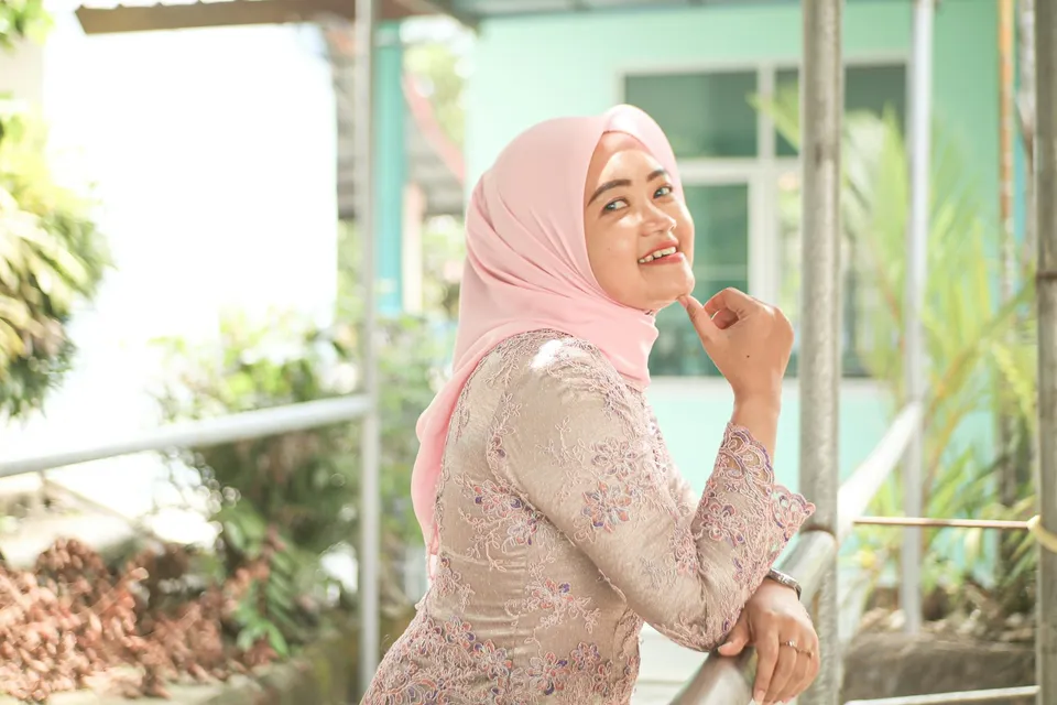 Seorang ibu berhijab merah muda dan kebaya brokat tersenyum sambil bersandar di pagar
