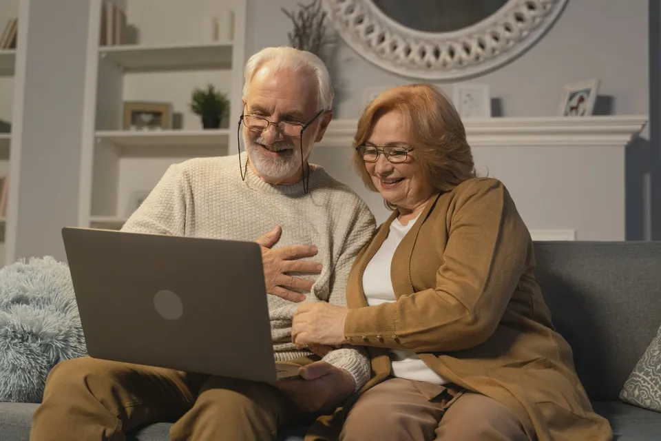 An older couple on the couch smiles at a video call on their laptop, one hand on his chest