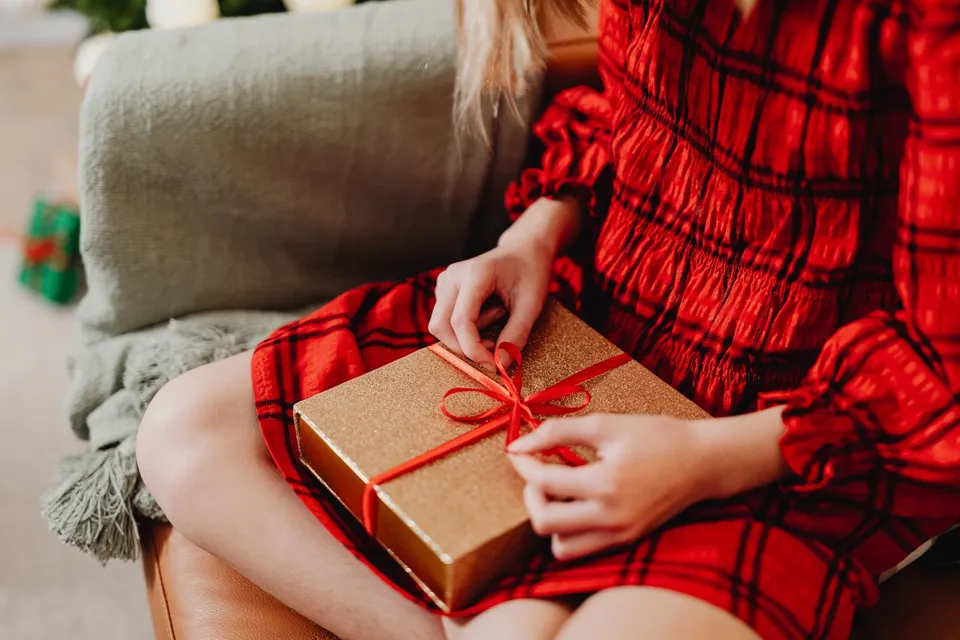 Una mujer con vestido de cuadros rojos desata el lazo de una caja de regalo dorada en el sofá