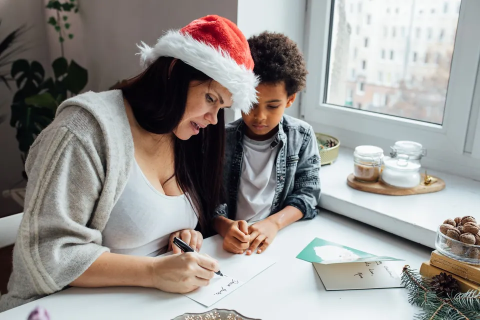 Une femme en bonnet de Père Noël et un garçon écrivent des cartes de Noël à la main près d'une fenêtre