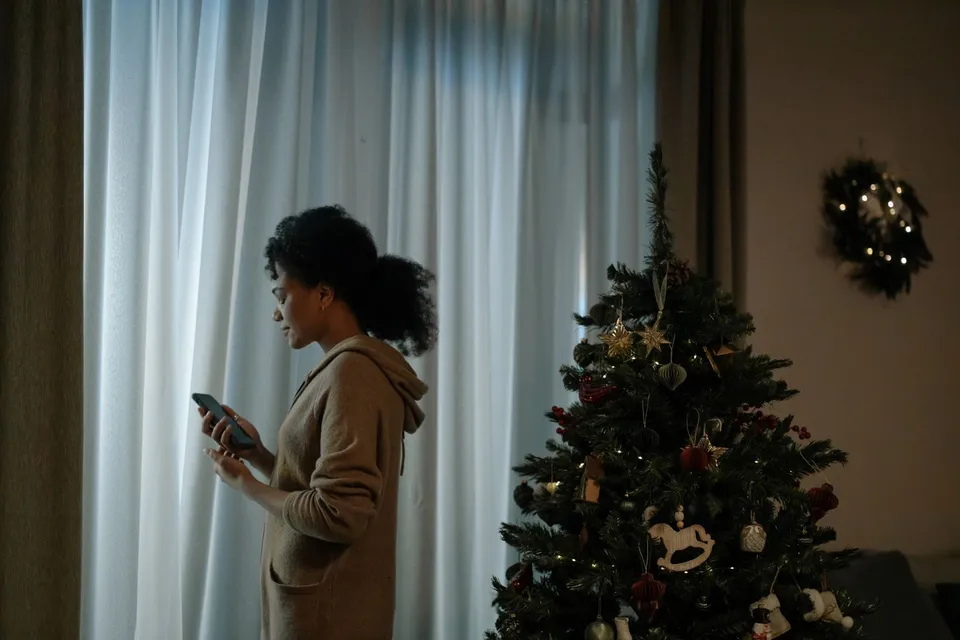 Une jeune femme tient son téléphone près d'un sapin de Noël décoré, devant une fenêtre