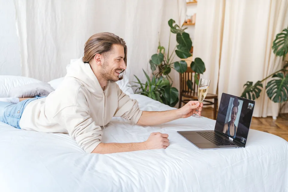 Um homem deitado na cama sorri e brinda com uma taça diante do notebook, em videochamada com uma mulher