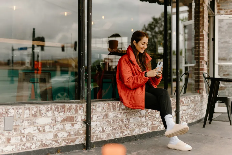Uma mulher de casaco vermelho sentada na janela de um café sorri enquanto lê o celular