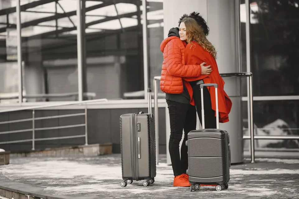 A couple in red coats hugging outside an airport, with two suitcases next to them