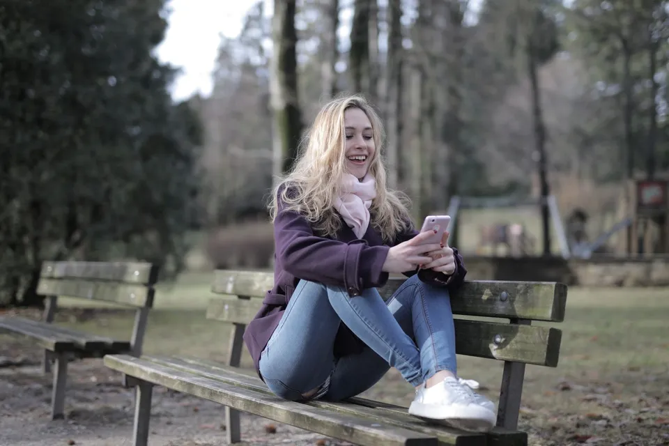 A young woman sitting on a park bench in winter, smiling at her phone