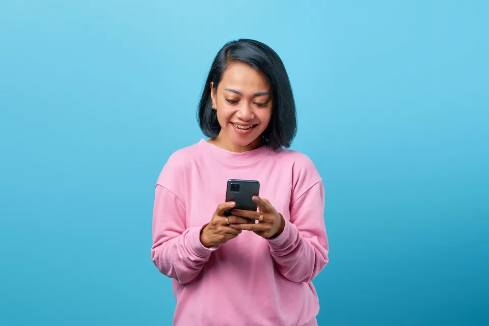 Seorang perempuan berbaju merah muda tersenyum melihat layar ponselnya