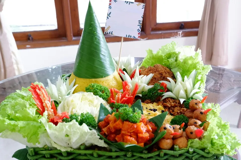 Tumpeng nasi kuning berbalut daun pisang dengan lauk di sekelilingnya dan kartu ucapan kosong di atasnya