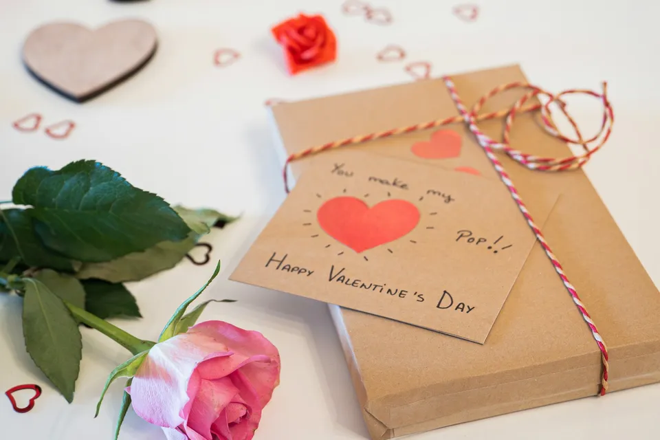 Une carte de Saint-Valentin écrite à la main avec un cœur rouge, posée sur un paquet en papier kraft à côté d'une rose rose