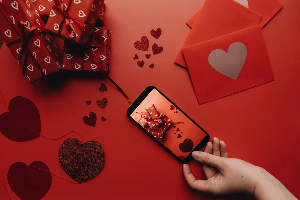 Une main touche l'écran d'un téléphone posé sur une table rouge, entre des cadeaux emballés et des enveloppes à cœur